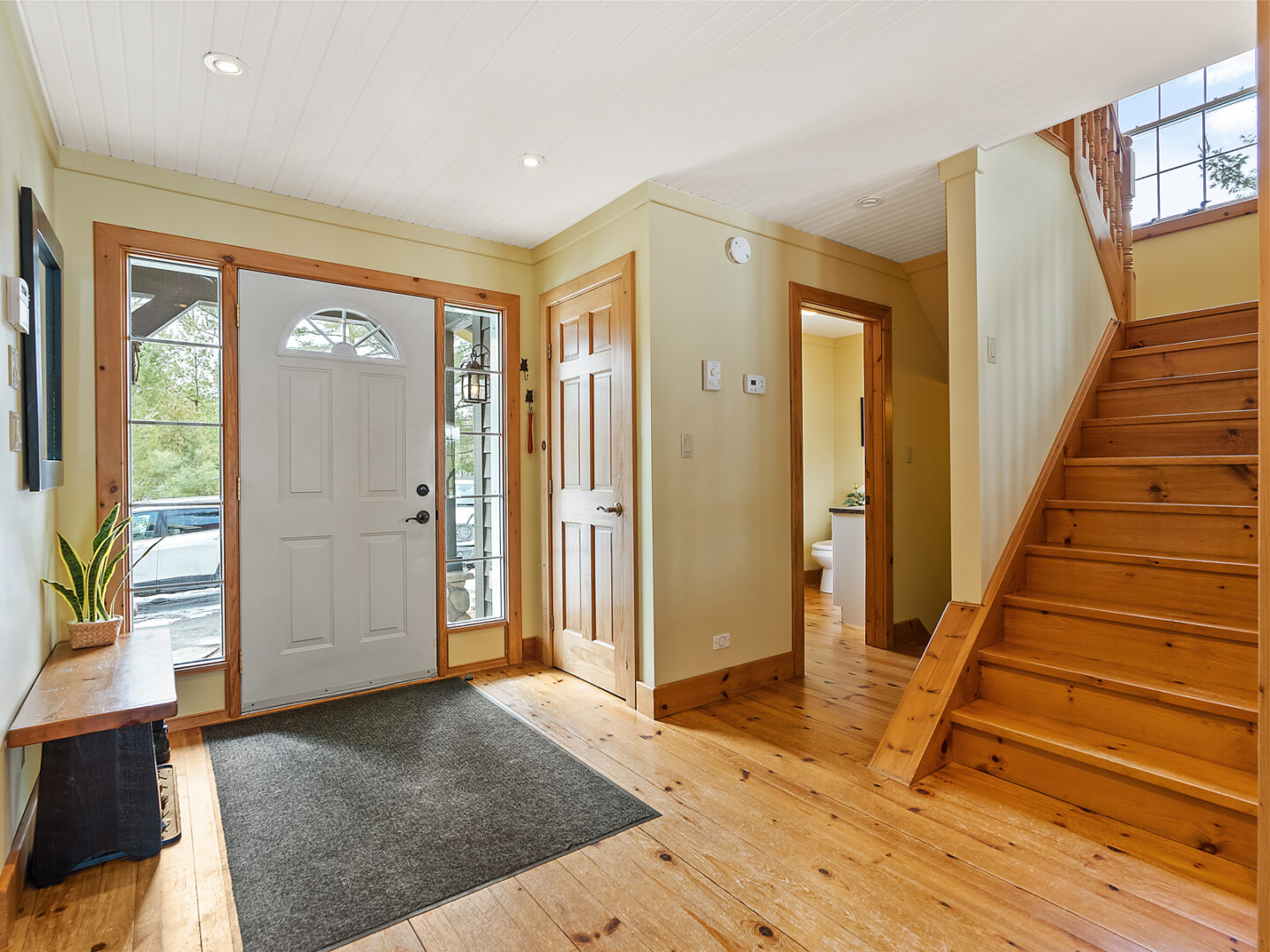 Main entrance of a lakefront home. Tall windows flank the front door, letting lots of light into the wide space, which has a bathroom off to the side and hardwood floors.