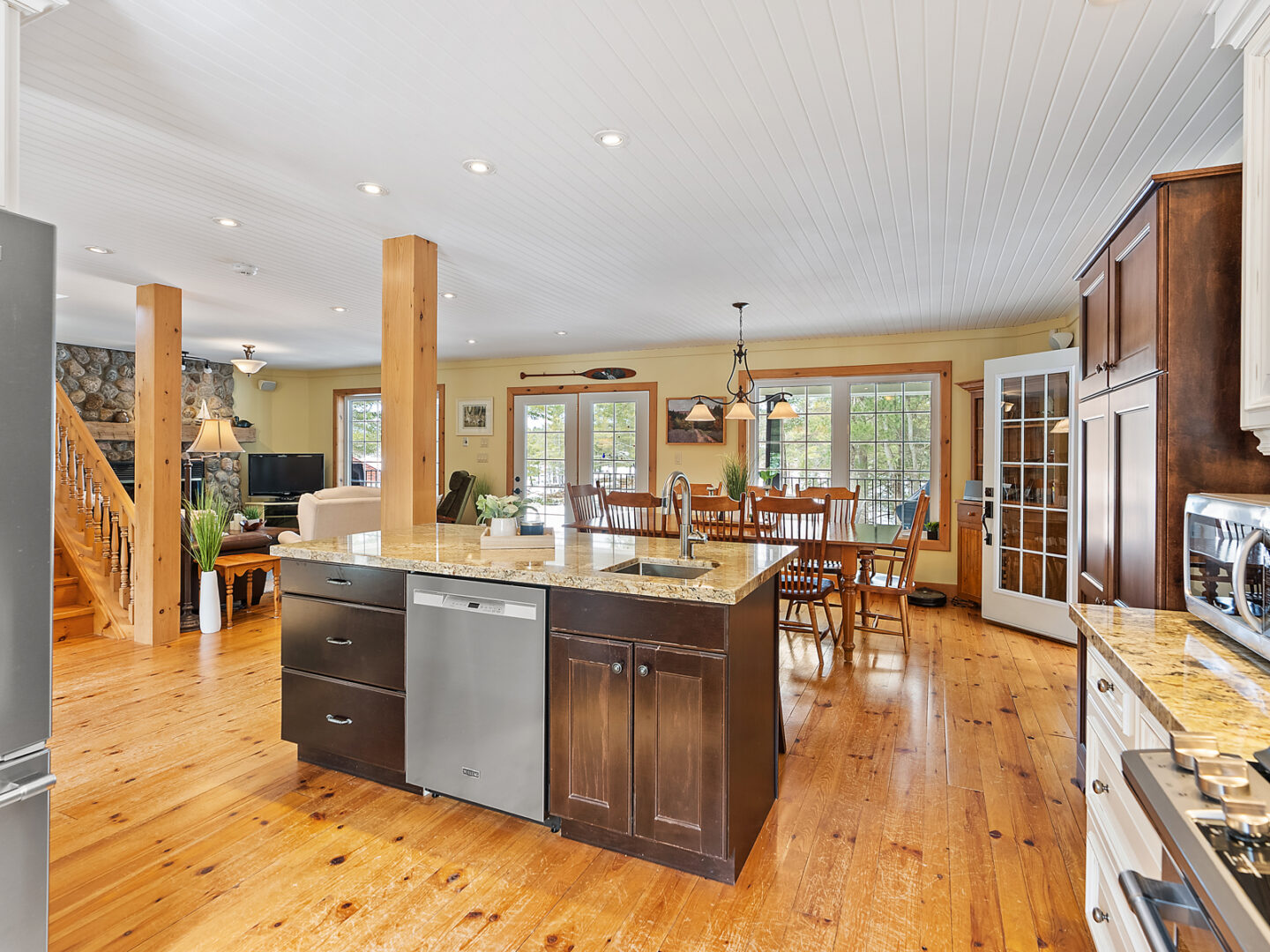 An open-concept kitchen space with warm hardwood floors and dark wood cabinets. A large island sits in front of a dining room table.
