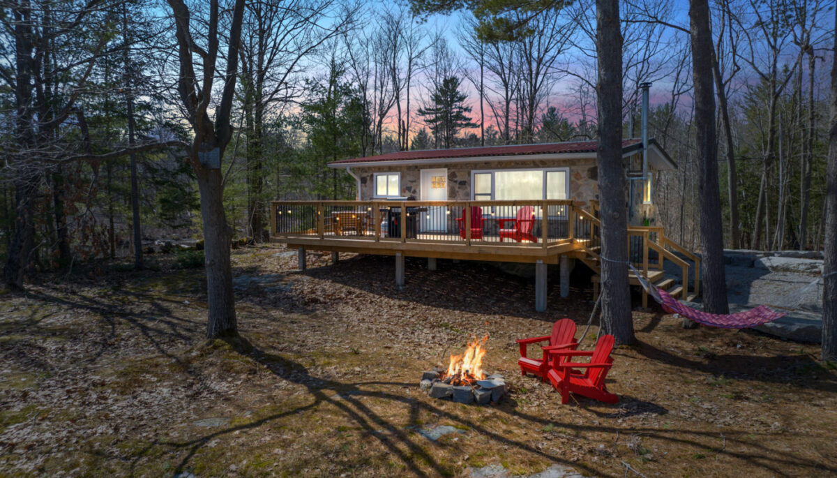 Outdoor space of a small cottage that has large, lit-up windows and a big back deck. There are trees all around, and two red Muskoka chairs sit in front of a small lit firepit.