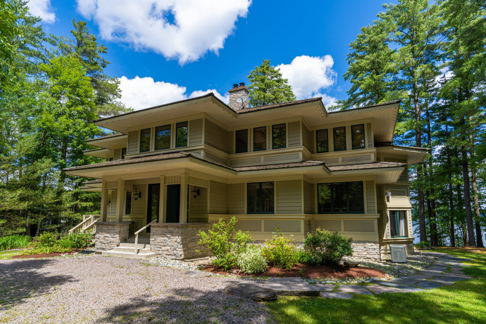 A large, luxury cottage with a large driveway and lots of windows, surrounded by green trees.