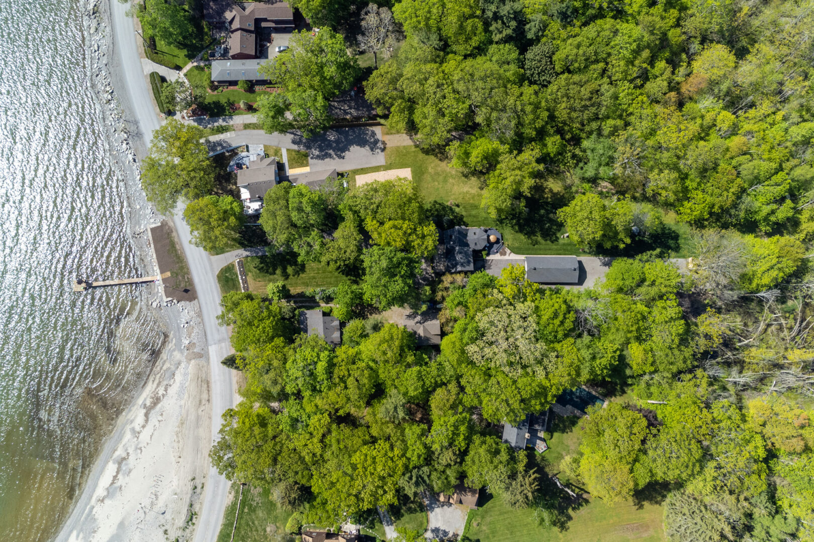 Overhead view of a large cottage lot. A big cottage with a detached garage is surrounded by trees. A long dock extends into the lake off the shoreline..