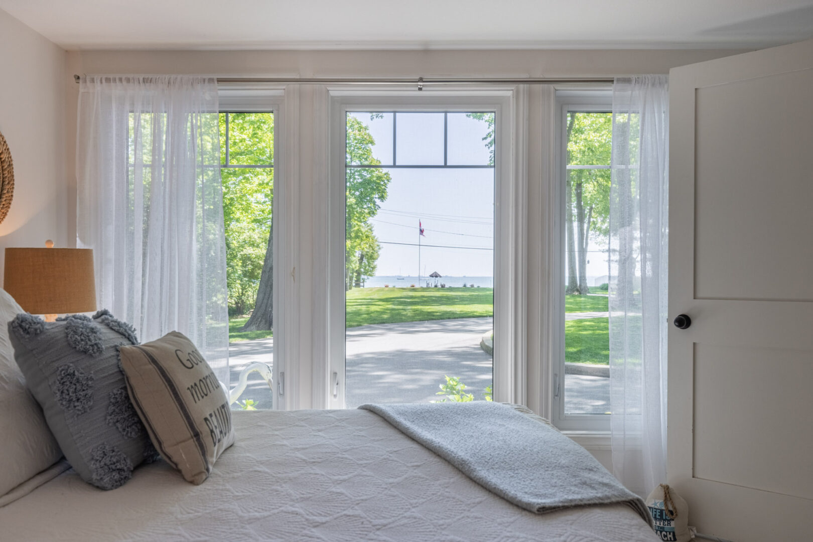 View across the bed and out the large windows of a bright bedroom. Grass and a lake can be seen out the window.