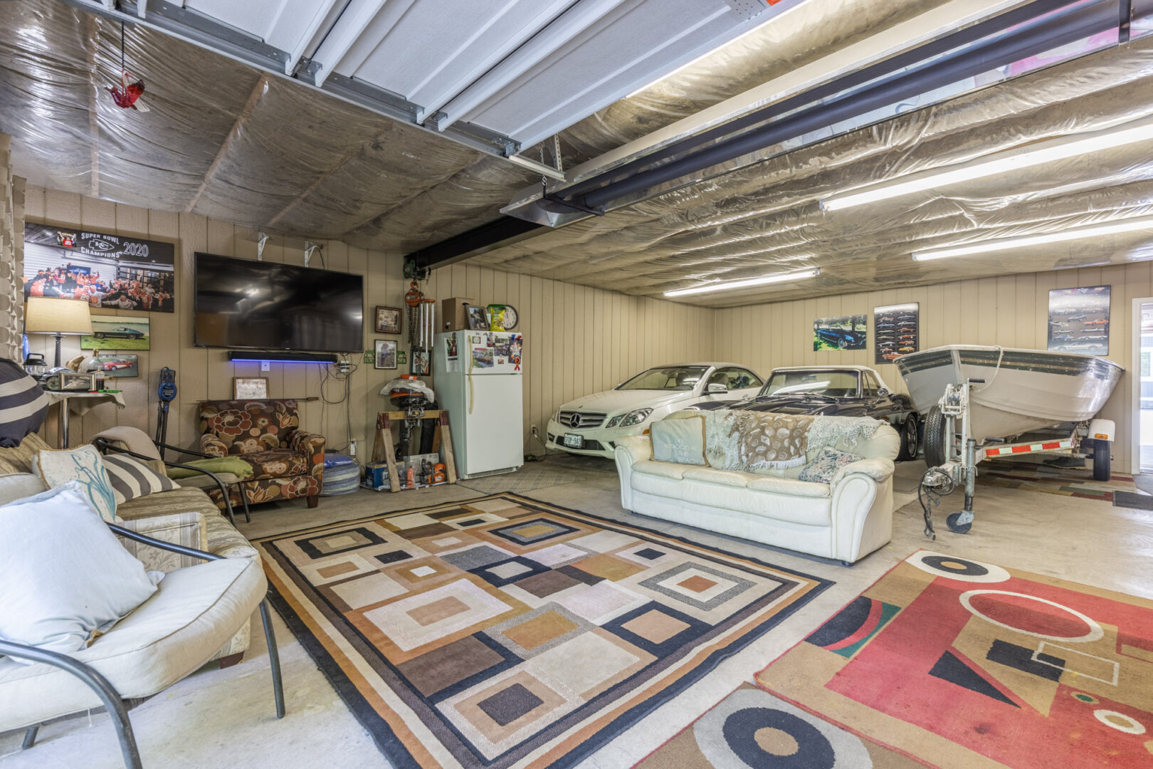 A large, insulated garage area with couches, a television, and area rugs. Behind a couch, there are parked cars and a boat.