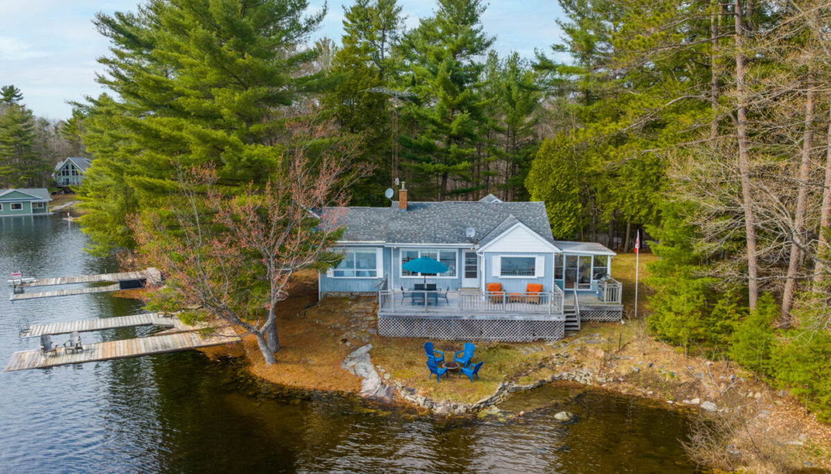 A cottage sits on the shoreline of a lake, surrounded by trees.