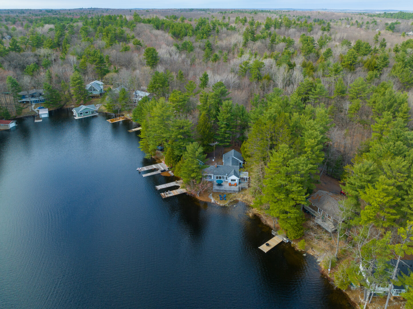 Overhead view of a cottage that sits on the shoreline of a still, blue lake, surrounded by green trees.