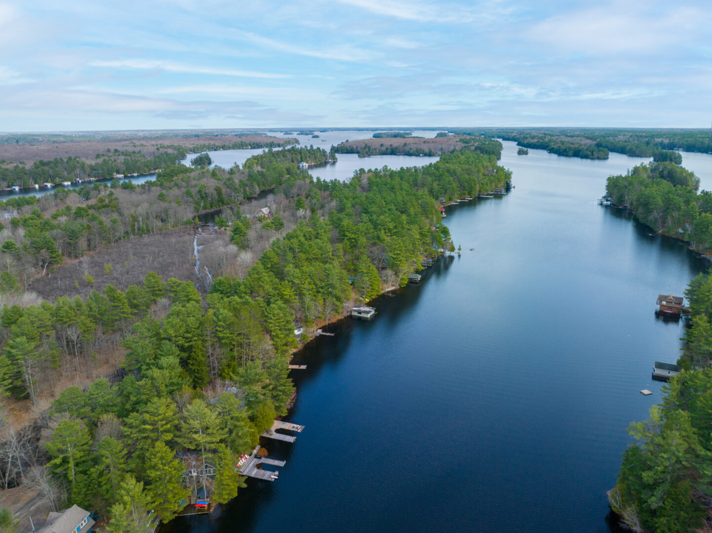 A narrow stretch of still, blue lake. Cottages and green trees line the shore on either side.