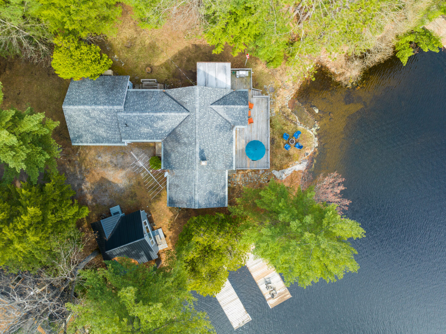 Aerial view of a cottage, which sits on the shore of a lake and is surrounded by green trees.