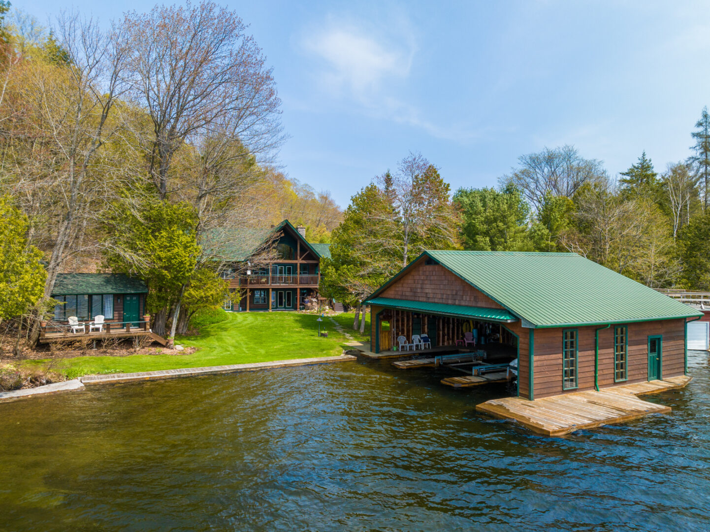 A big boathouse with a green roof sits in a lake, and up on the shoreline is a large cottage and a small guest bunkie.