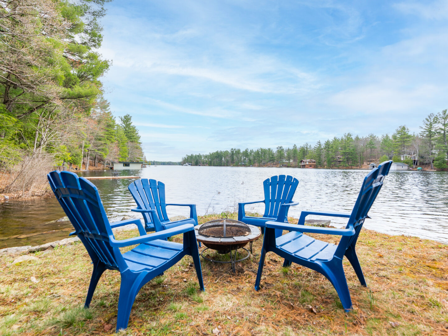 Four blue Muskoka-style chairs sit around a fire pit, lakeside.