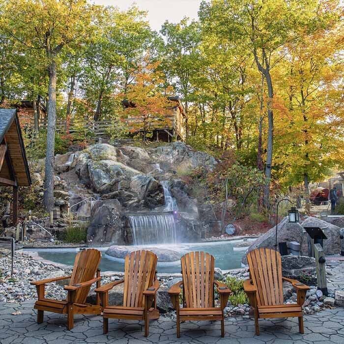 Four Muskoka chairs in front of Spa Nordik waterfall and pool with forest in the background