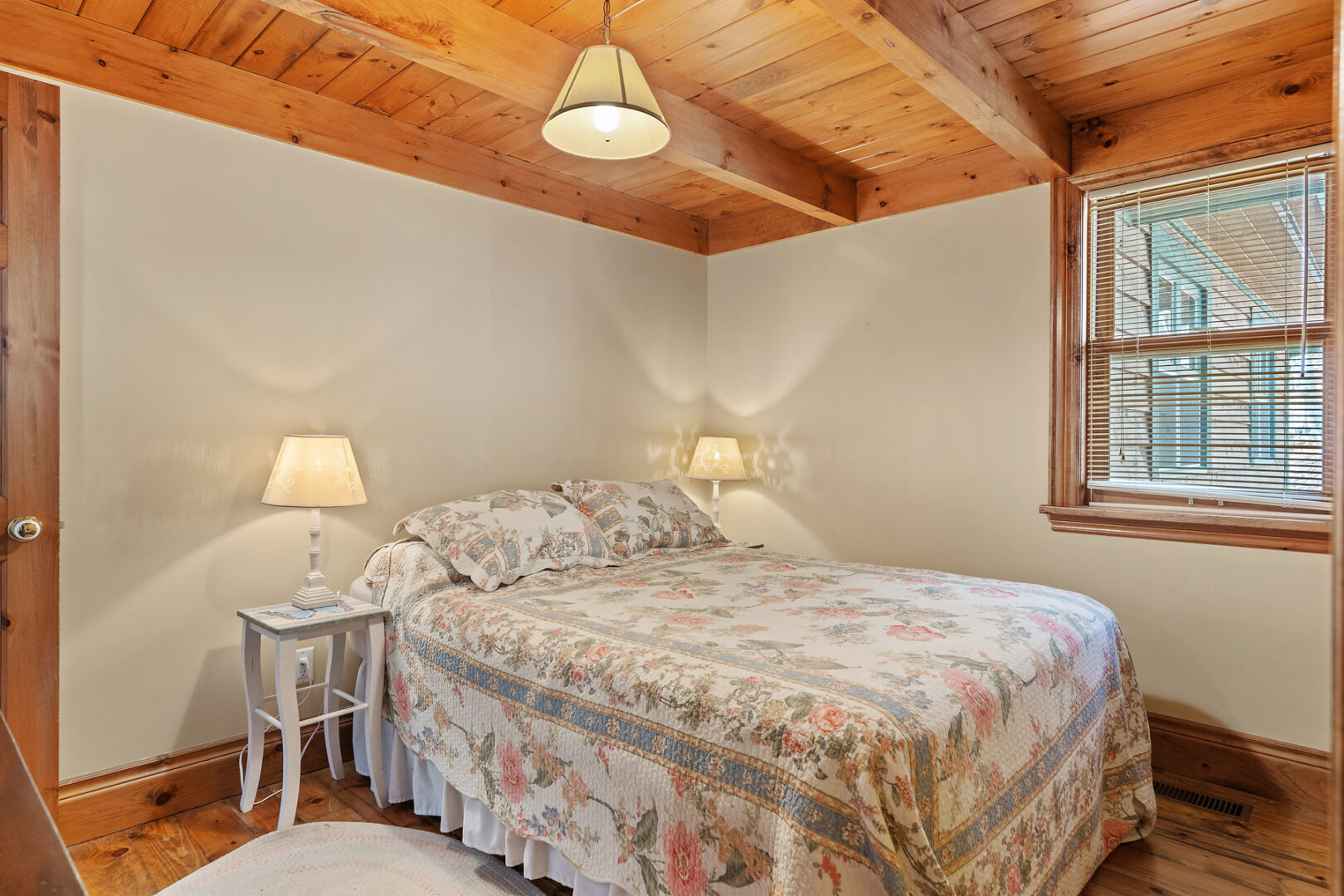 A small bedroom with a wood-panelled ceiling, white walls, and a floral bedspread.