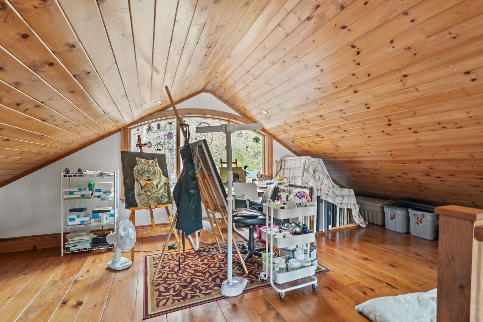 A large loft space with slanted wood-panelled ceilings and a big window, set up like an art room with easels and painting supplies.