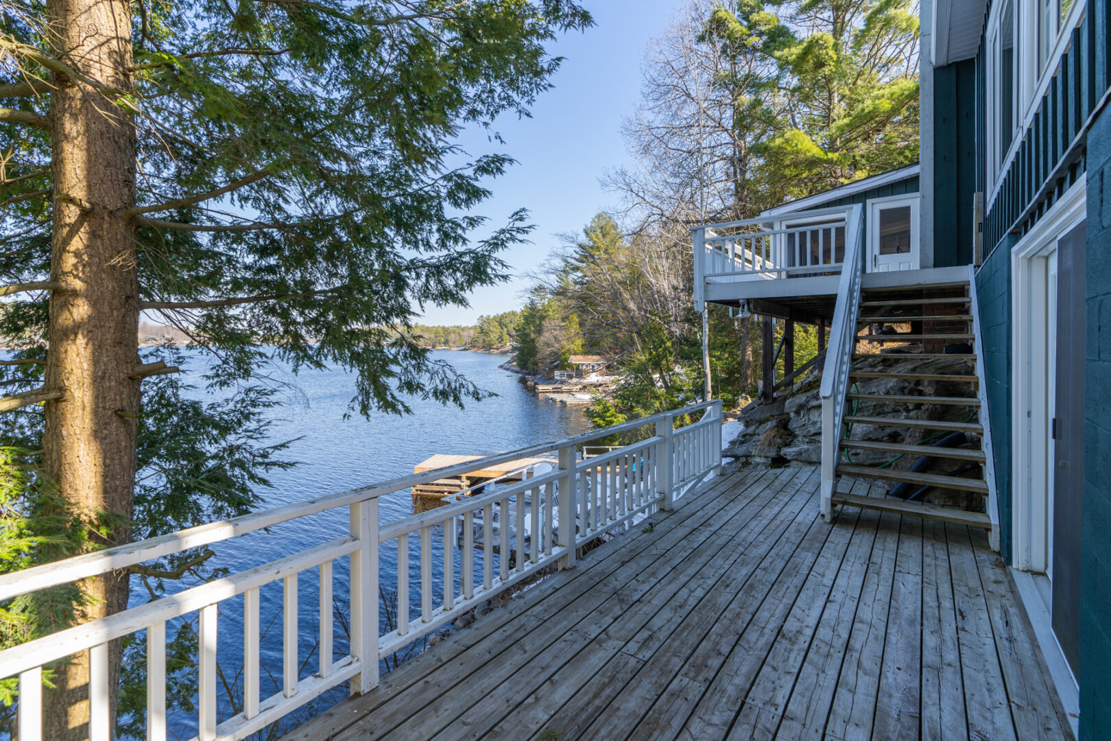 A large deck with a white railing extends off the side of a cottage, overlooking a lake.