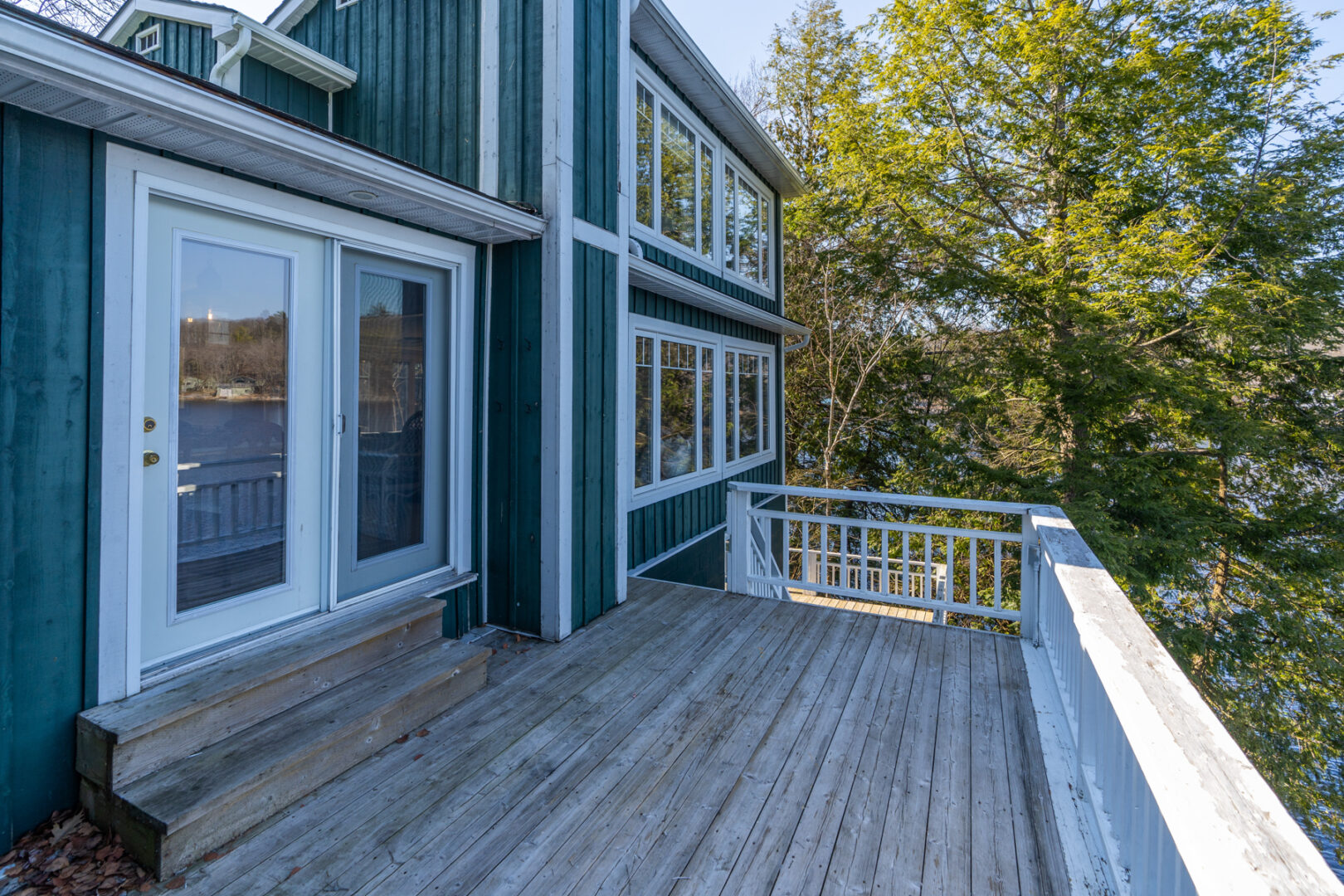 A large deck with a white railing extends off the side of a cottage, which has lots of windows.