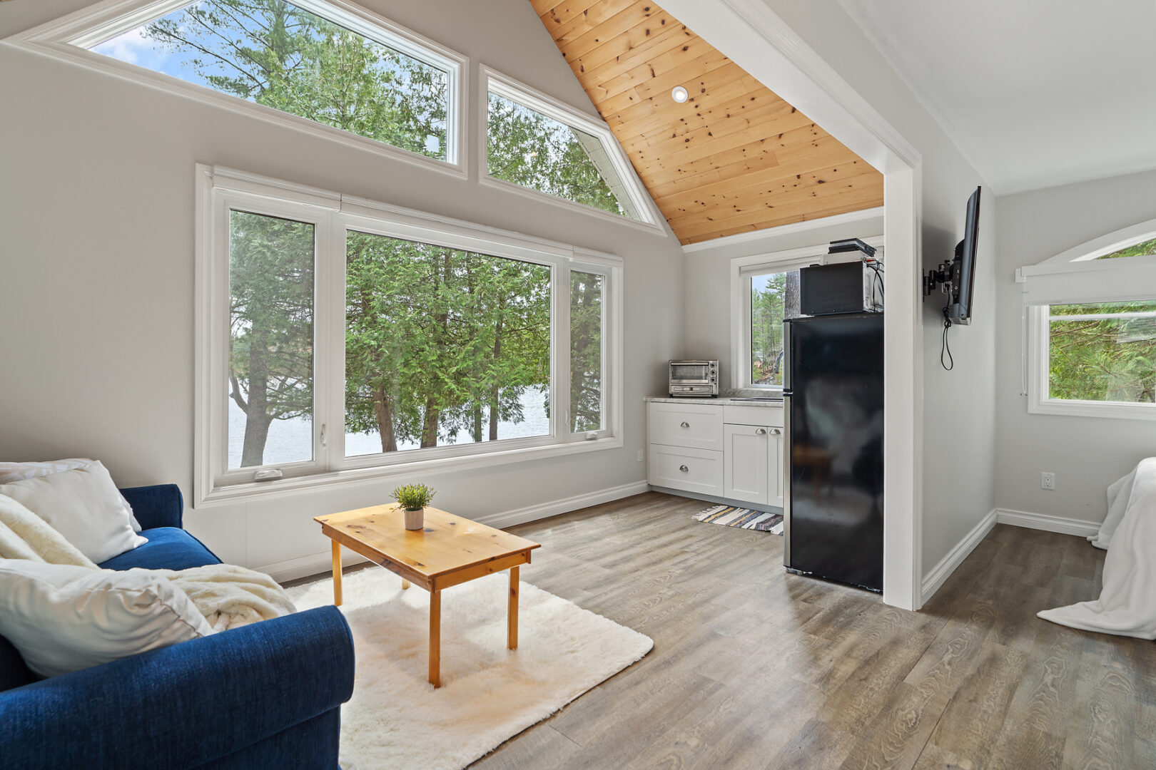 A bright, open-concept but small bunkie living area with a couch, a kitchenette, large windows and a wood-panelled ceiling.