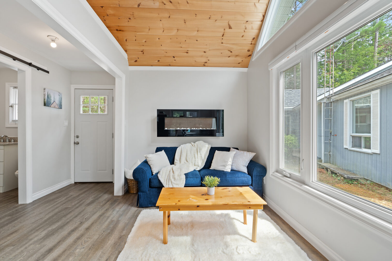 A small, bright bunkie living area with a couch, large windows, and a wood-panelled ceiling.