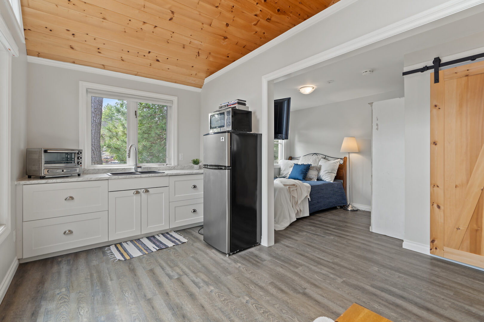 A studio-style bunkie living space with a kitchenette, a bed, and a wood-panelled ceiling.
