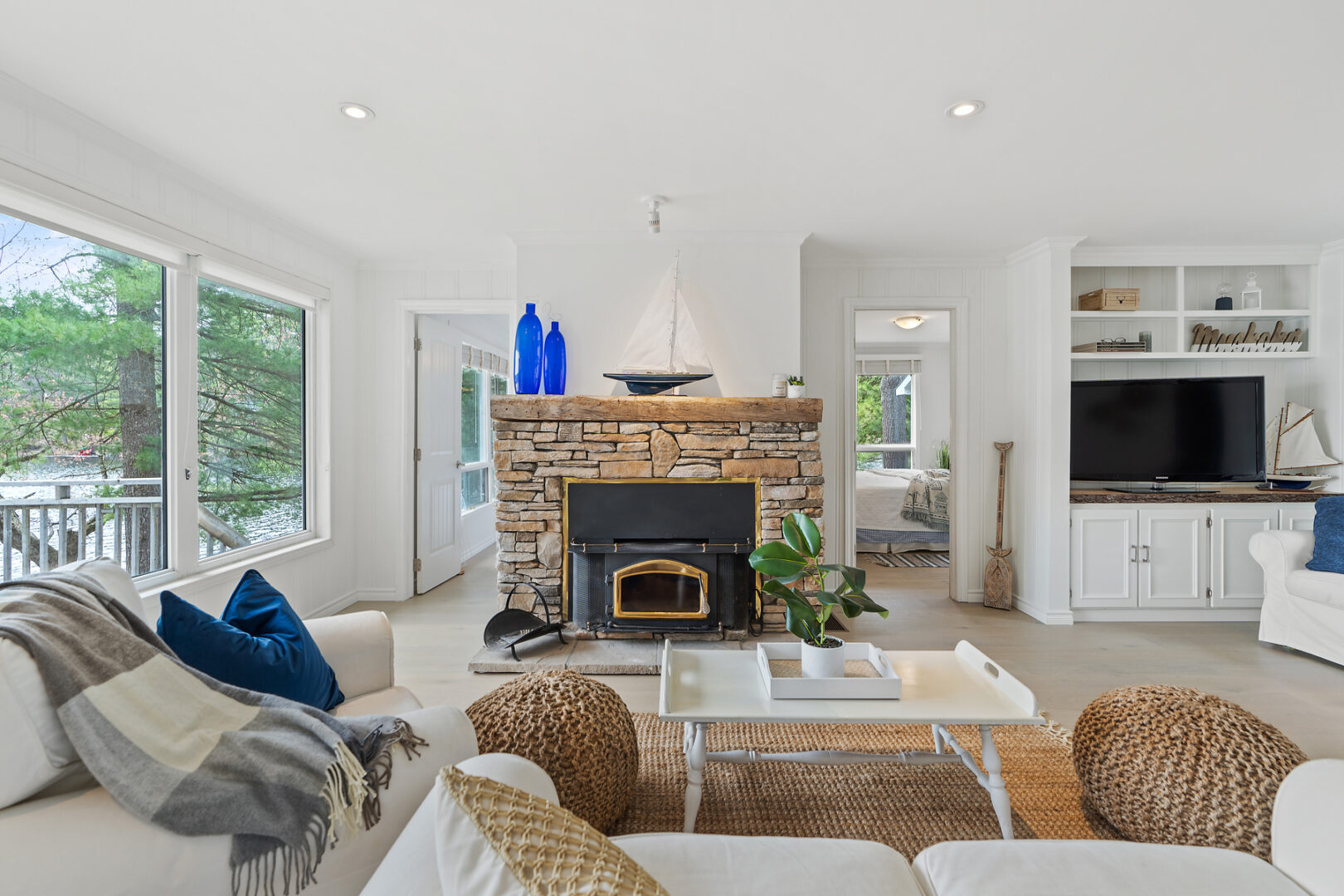 A bright, open cottage living space with a fireplace, couches, large windows, and white walls. Two doorways lead to bedrooms off the living area.
