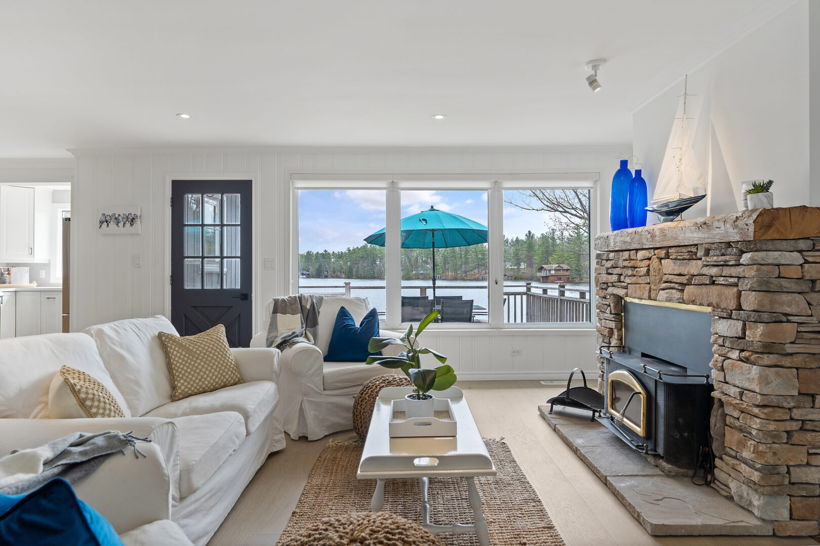 A bright cottage living space with a fireplace, couches, hardwood floors, and large windows looking out to the water.