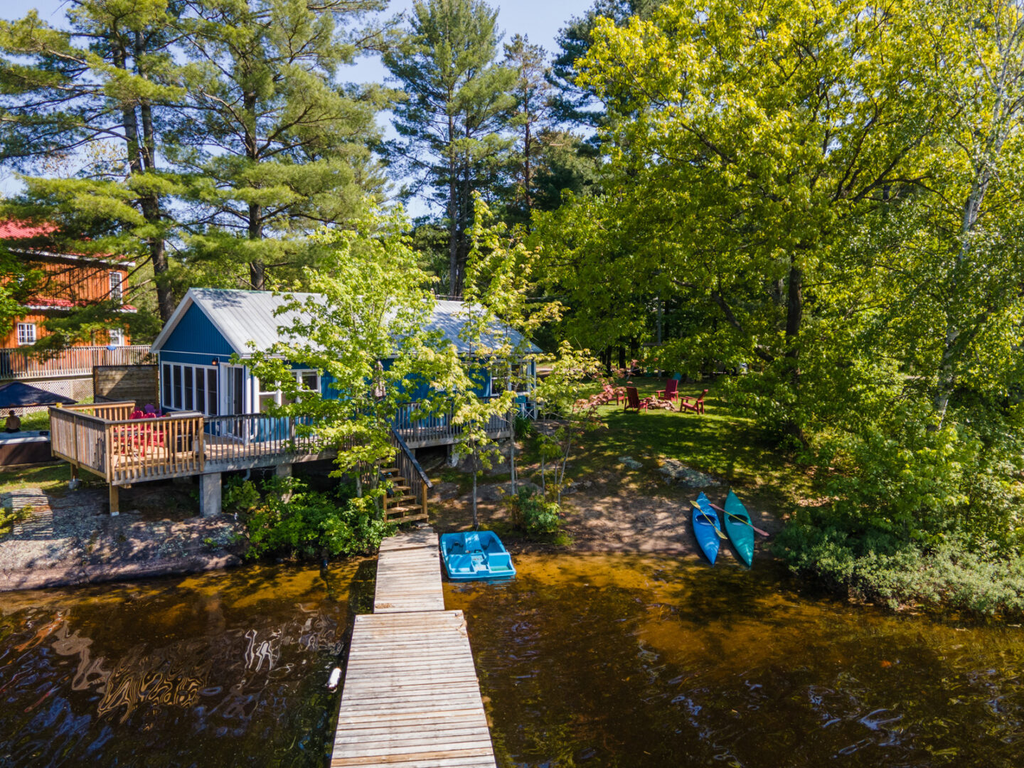 A long dock stretches into a still lake, from a sandy shoreline. A small blue cottage shrouded by green trees sits just up the shore.