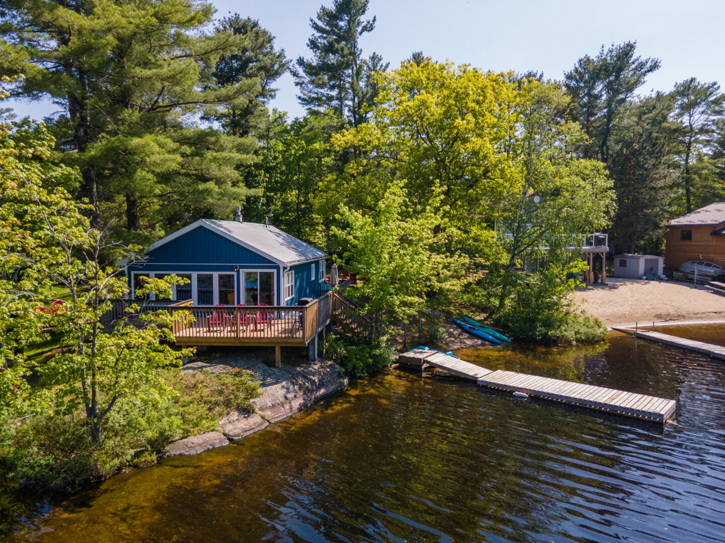 A small blue cottage with a big deck, a sandy shore, and a long dock sits lakeside. Green trees surround the land behind..