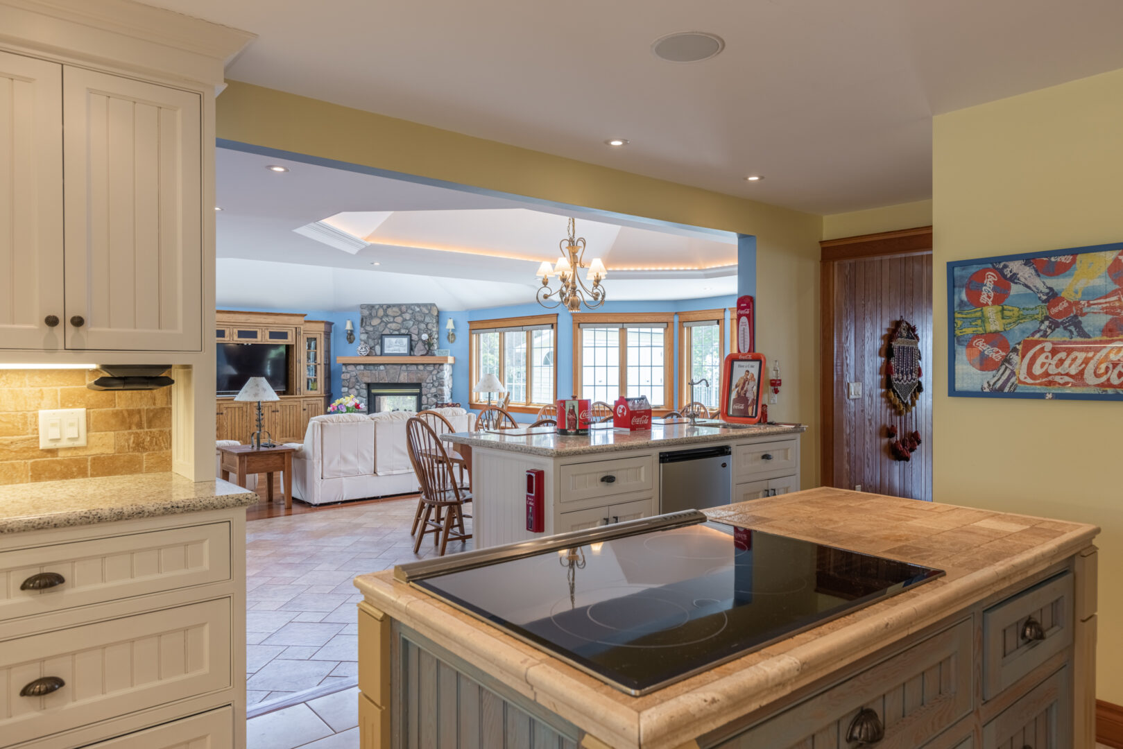 A large kitchen island with a stovetop sits in the middle of a kitchen area that leads into a bright living space.