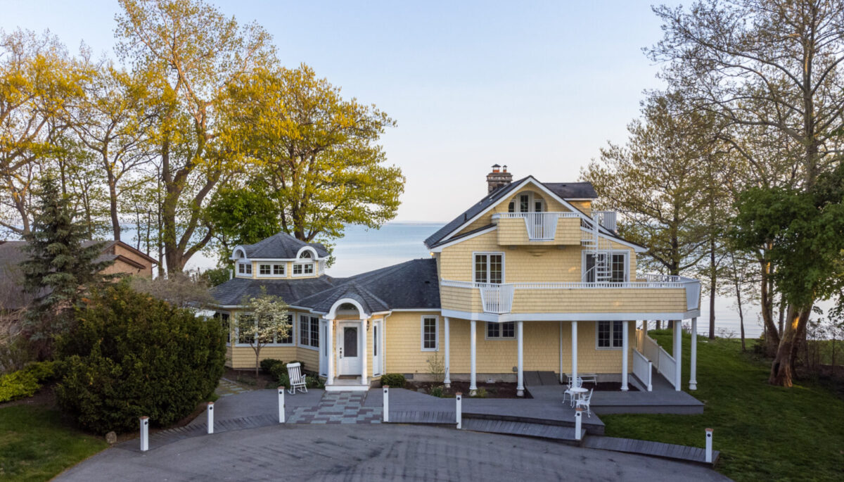 Front exterior of a large yellow cottage with white balconies and a paved driveway.