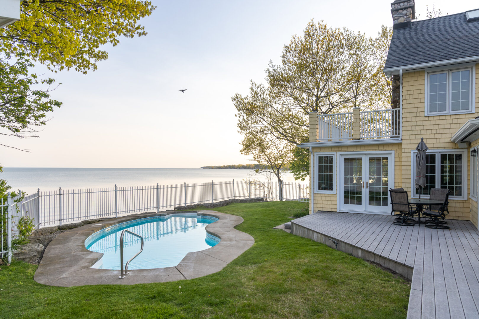 Back lawn area of a lakefront house, with an in-ground pool surrounded by a white fence overlooking the lake.