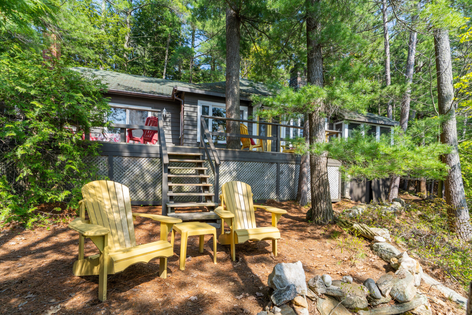 Two yellow Muskoka chairs sit in front of stairs leading up to a small cottage with a large deck.