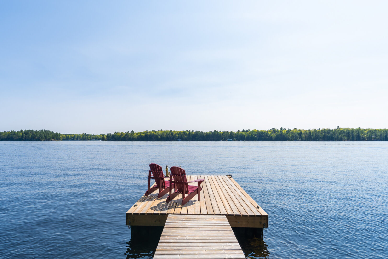 A long dock with two red Muskoka chairs on the end extends into a wide blue lake.