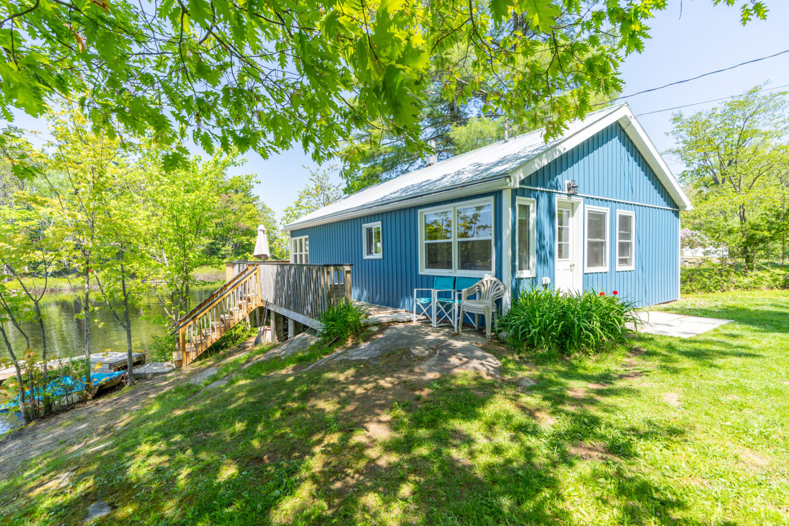 A small blue cottage sits on a grassy hill. A wooden deck stretches down the side, with stairs leading down to a lakeside dock.
