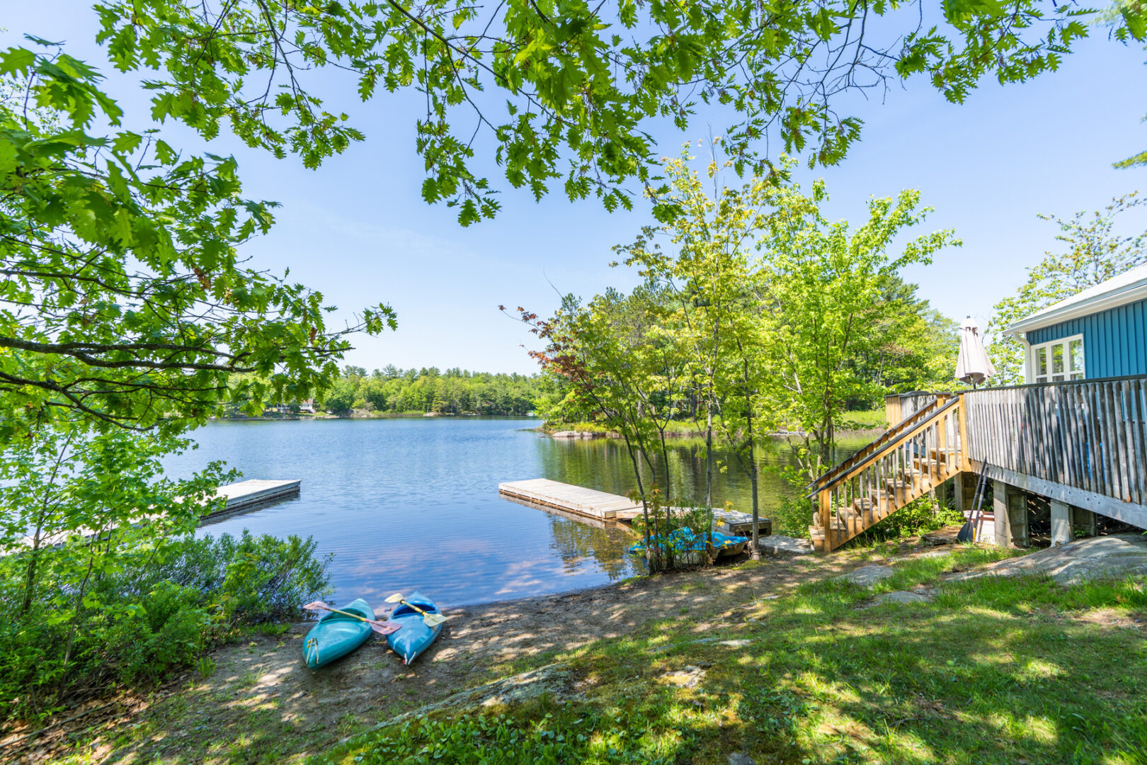 A shady lakeside area with a small sandy beach and a long dock off a grassy shore.