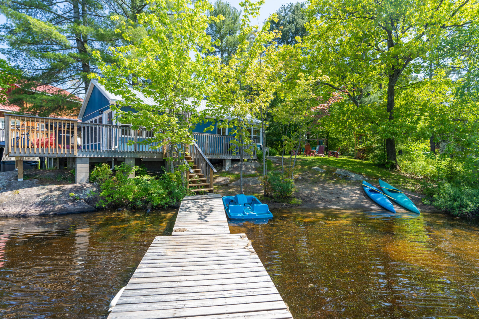 A long dock stretches into a still lake, from a sandy shoreline. A small blue cottage shrouded by green trees sits just up the shore.