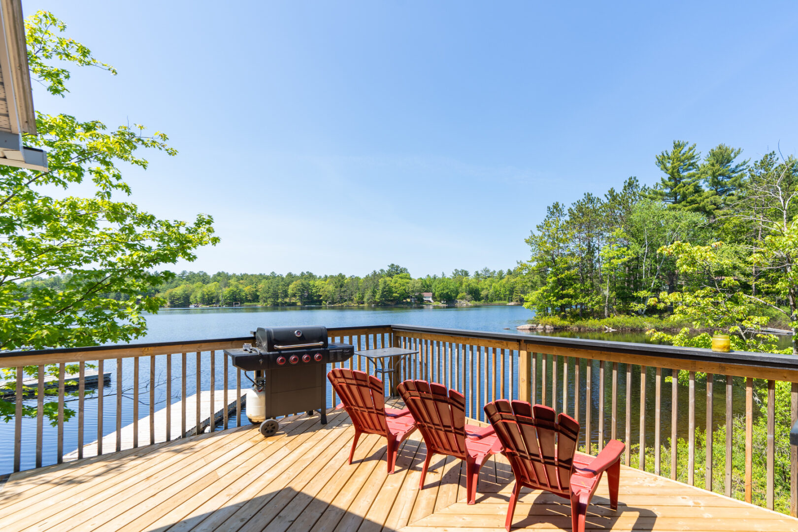 A large deck with a barbecue and three red Muskoka chairs looks out across a blue lake.