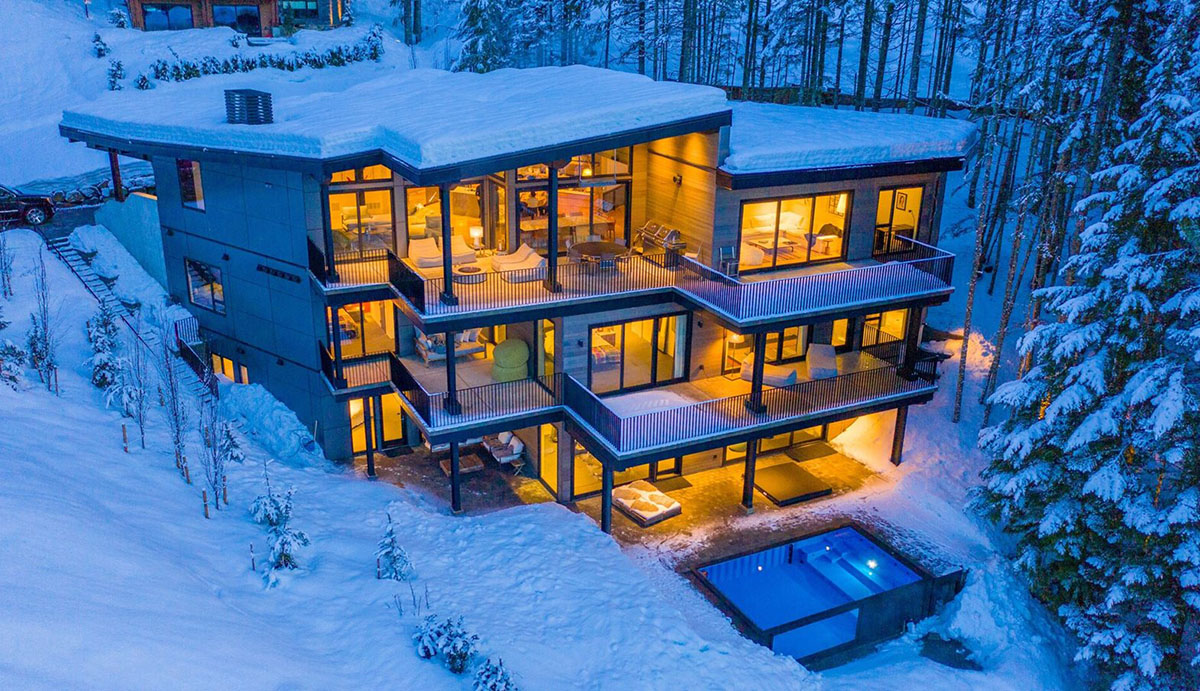 Chalet with lots of windows is illuminated from the inside against the snow from outside at dusk