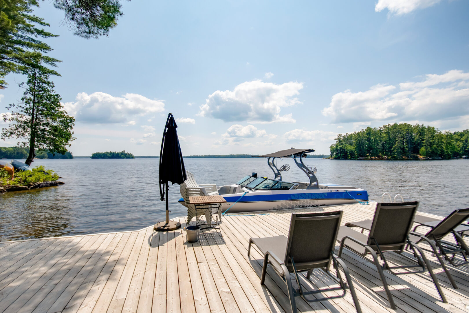 View over a lake from a big dock, which has lounger chairs and a closed umbrella. A boat sits in the water by the dock.