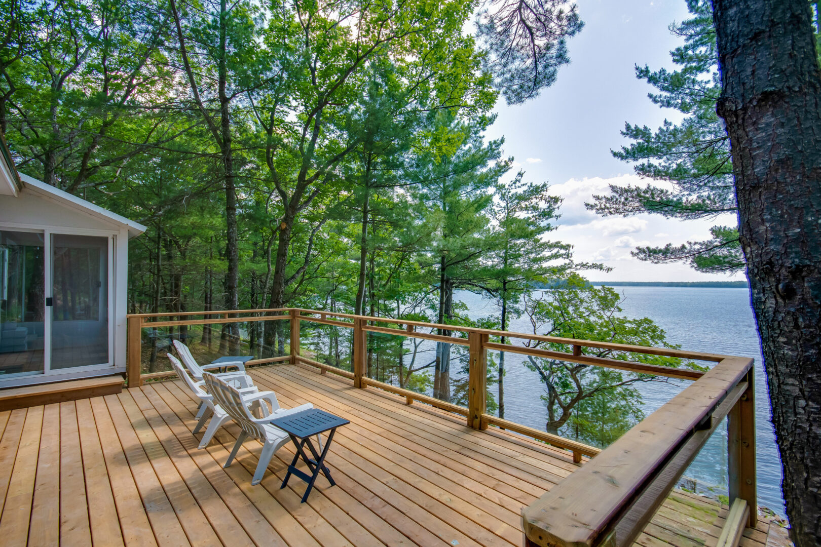 A spacious wooden deck with three chairs and a glass railing looks out over a lake.