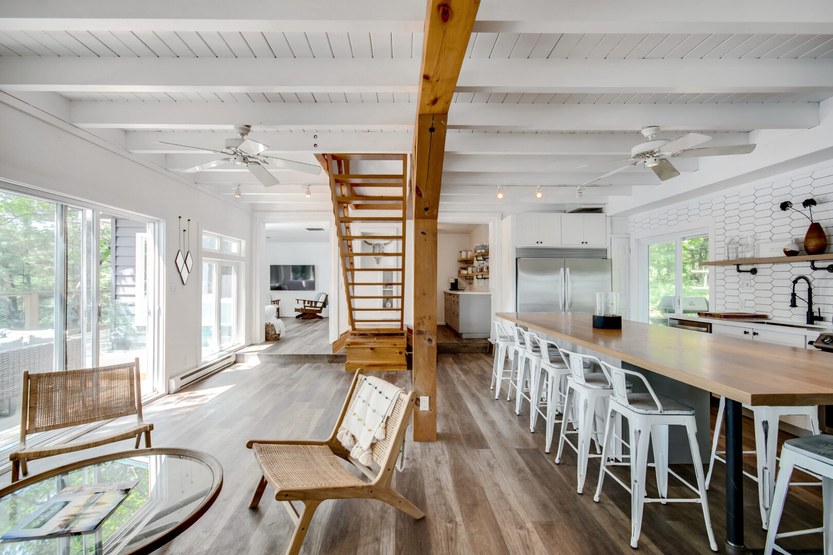 A bright kitchen and dining area with a table and chairs, island seating, and large windows on one wall. A hardwood staircase leads up to a second floor at the end of the room.