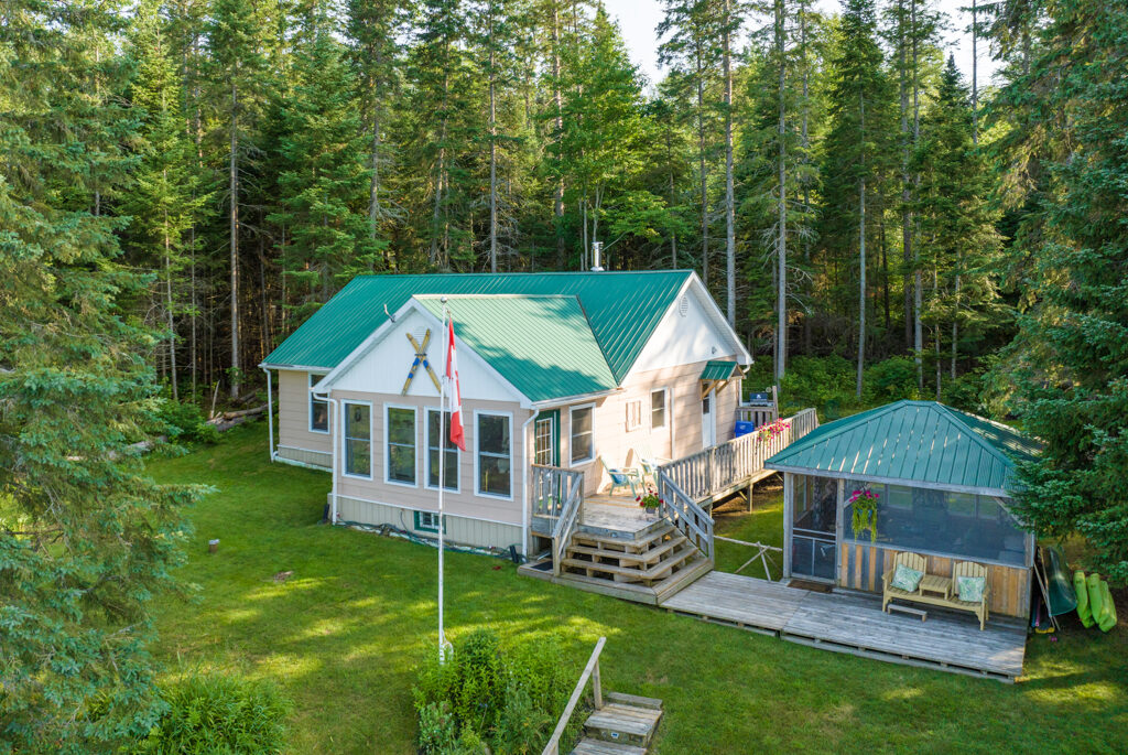 A cottage with big windows and a green roof sits on a grassy property. Beside it, there is a large, detached screened-in gazebo, also with a green roof.