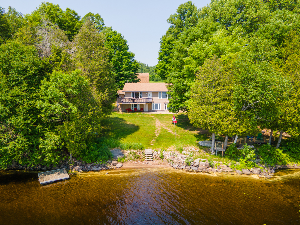A house sits up the hill from the sandy shoreline of a lake, surrounded by trees.