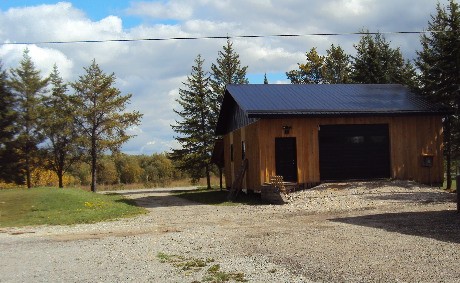 A dirt driveway of a rural property. A cedar garage with a black roof and door sit to the side of the road.