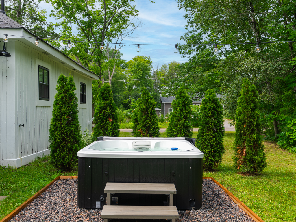 A hot tub sits beside a small house, behind a row of tall shrubs.