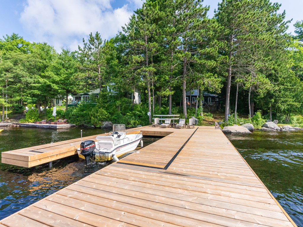 A big, new dock extends out into a lake. Four lounger chairs sit on the dock close to the shore, facing out toward the water.