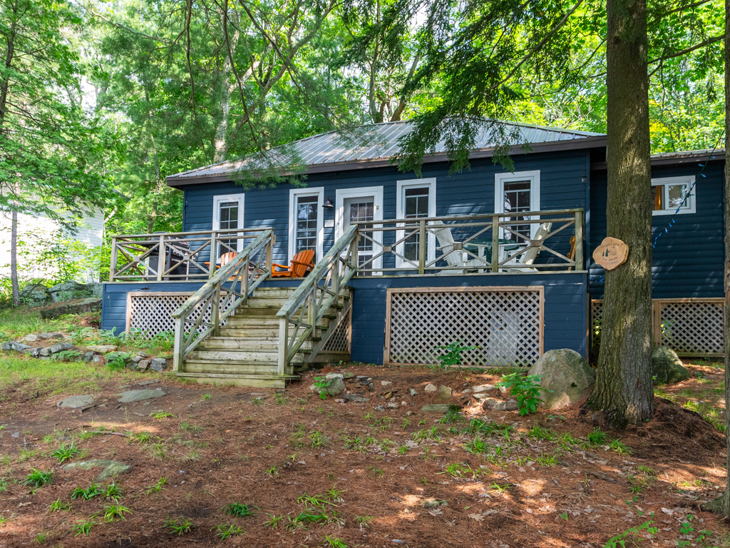 A bungalow cottage with a dark blue exterior sits surrounded by green trees.