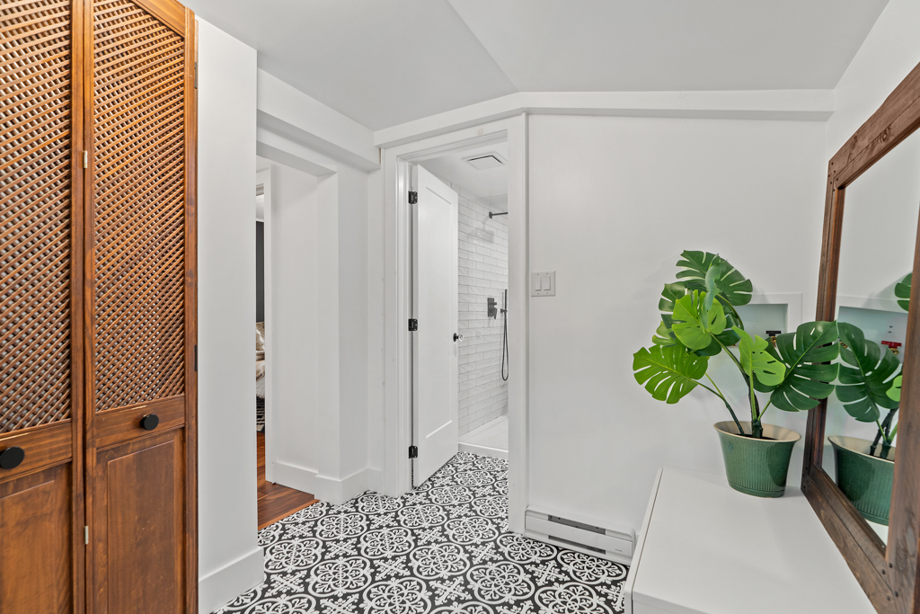 The end of a hallway that leads into a bathroom area, with black and white patterned tile on the floor and a green plant on a table.