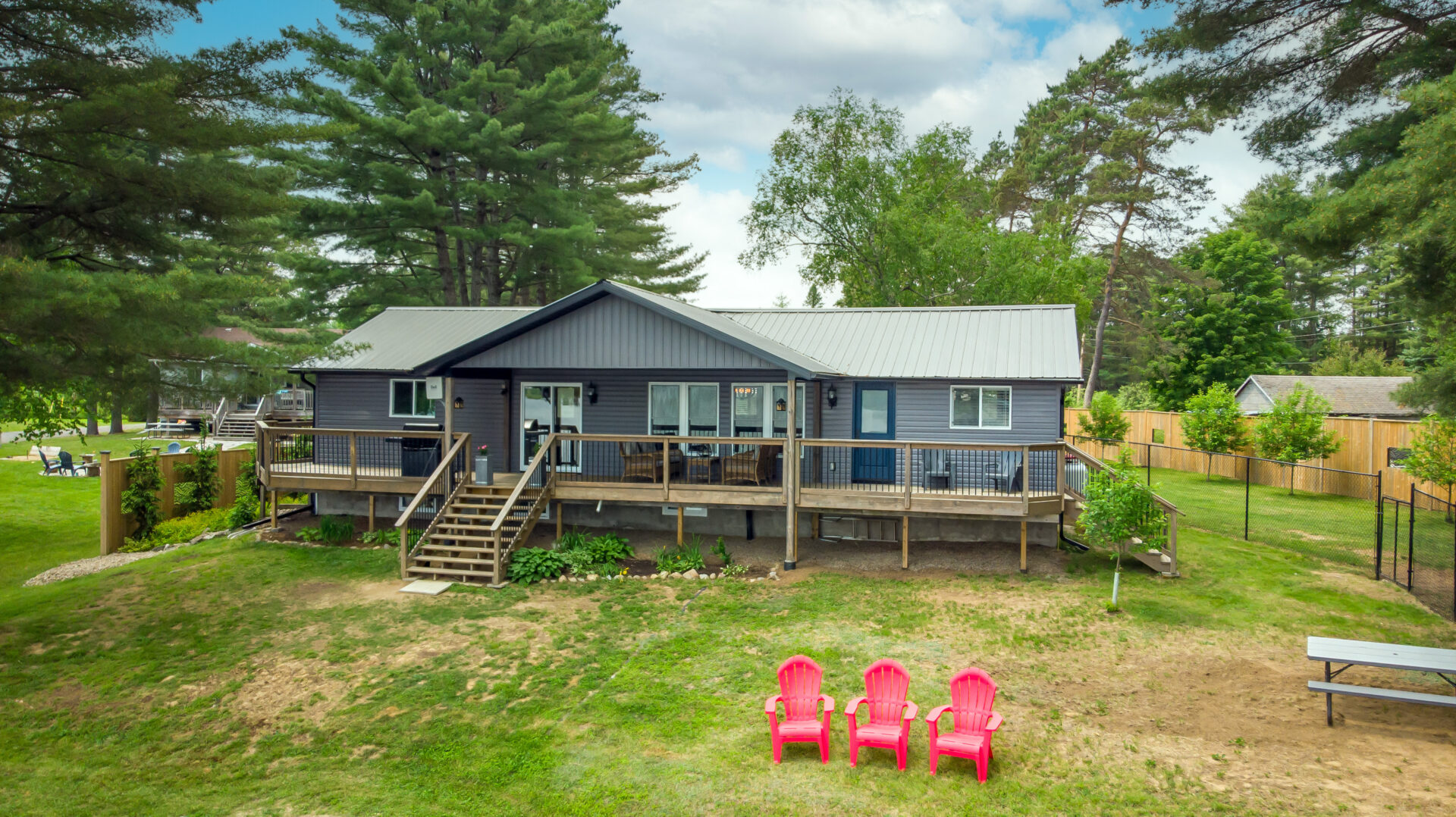 Three red Muskoka chairs sit on a sloping grassy yard in front of a bungalow cottage.