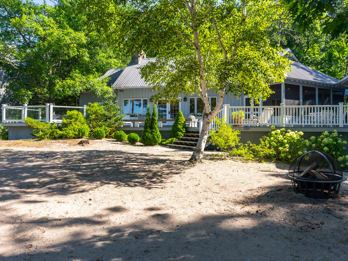 A wide, sandy area lined in greenery, in front of a large luxury cottage.