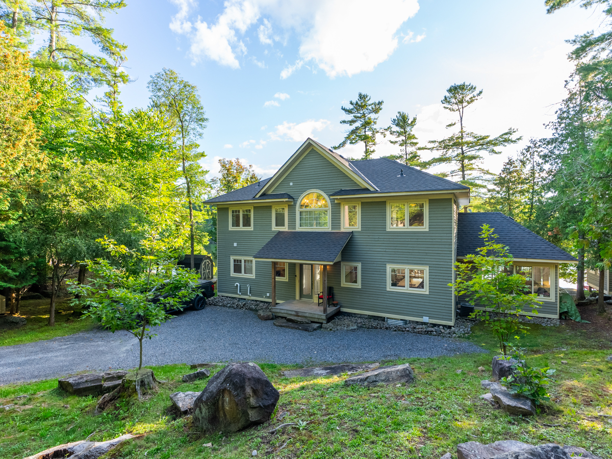 A large cottage with a big driveway sits surrounded by green trees.