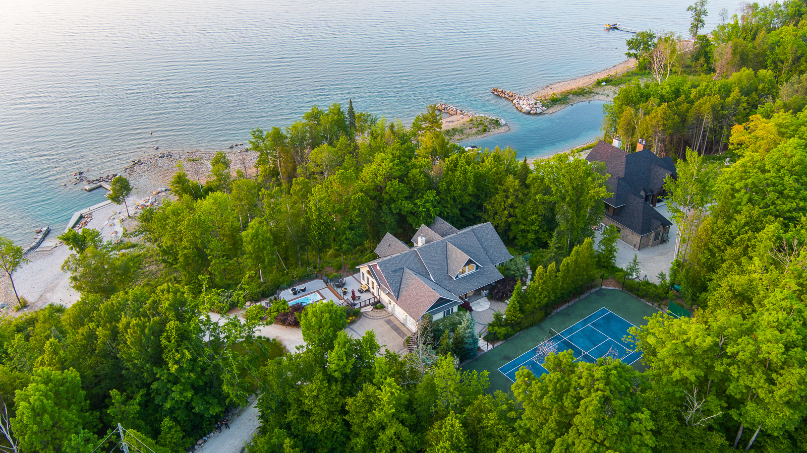 A large luxury property just off the waterfront with a big outdoor patio and a tennis court, surrounded by green trees.