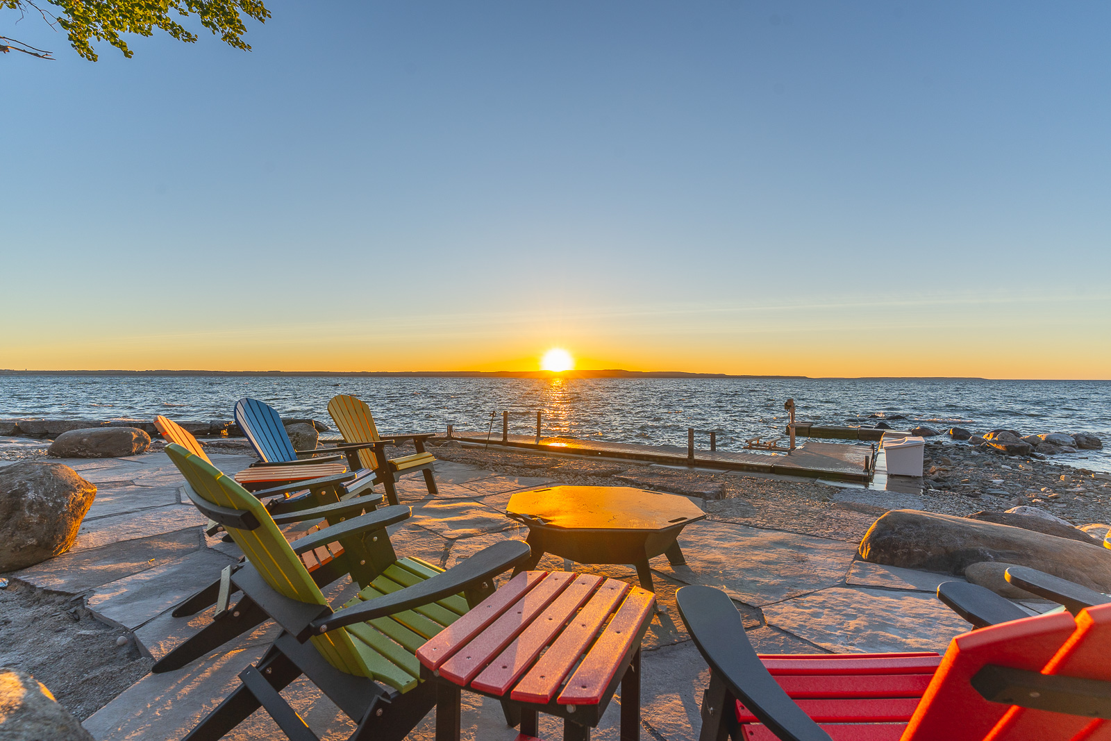 A large patio with a fire table, surrounded by colourful Muskoka chairs, looking out over the lake at sunset.